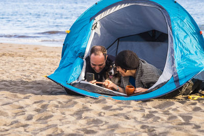 Smiling couple using mobile phone while lying in tent at beach
