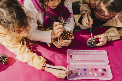 Girls using paintbrushes for coloring pine cones at picnic table in park on sunny day