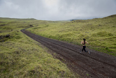 Rear view of woman jogging on dirt road amidst green landscape against cloudy sky