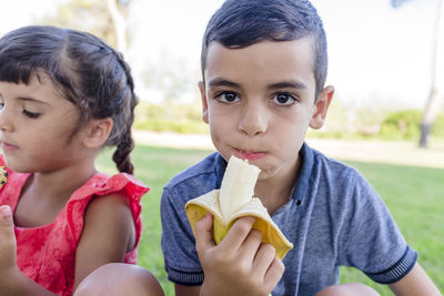 Two kids eating banana sitting outdoors on the grass in a park.