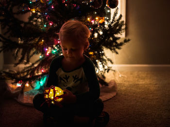 Boy holding illuminated decoration while sitting by christmas tree