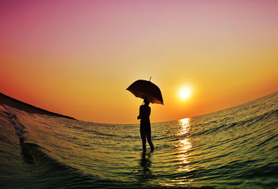 Silhouette woman with umbrella standing in sea against orange sky