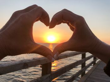 Cropped image of hands forming heart shape against sky during sunset