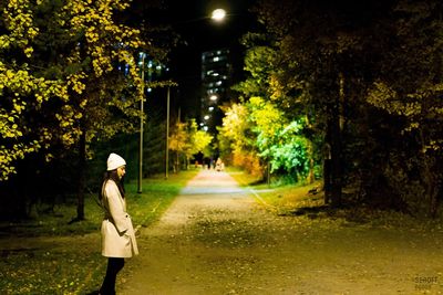 Side view of woman standing on footpath amidst trees at night