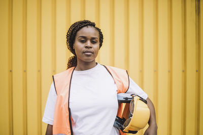 Portrait of confident female construction worker with hardhat against yellow metal wall