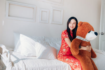 Young woman in traditional clothing holding teddy bear while sitting on bed at home