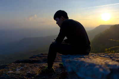 Man sitting on rock against sky during sunset