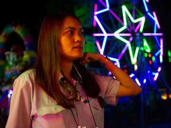 Woman looking away while standing against illuminated ferris wheel at night