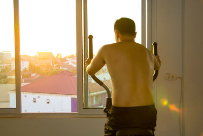 Rear view of shirtless man standing against window