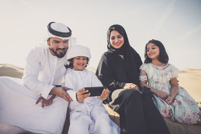 Arabian family taking selfie while sitting at desert against sky