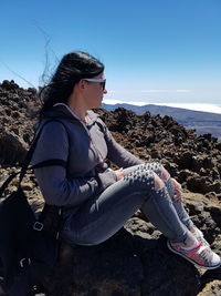 Side view of young woman sitting on rocky mountain against sky during sunny day