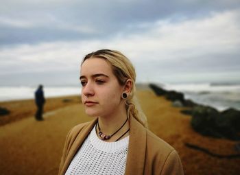 Thoughtful teenage girl looking away while standing at beach against cloudy sky