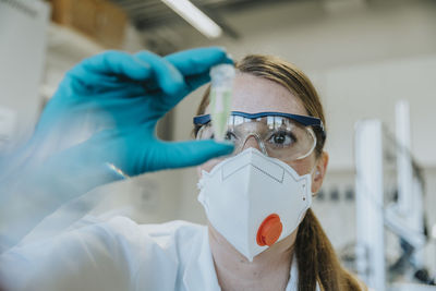 Young woman wearing protective face mask and eyeglasses examining test tube at laboratory
