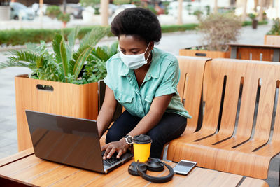 A young african girl in a medical face mask works with a laptop at a table in a cafe. 