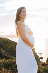 Side view of smiling pregnant woman standing at beach against sky