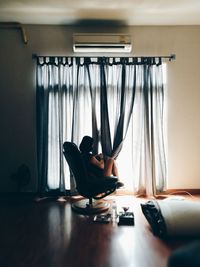 Woman playing with curtain while sitting on chair against window at home