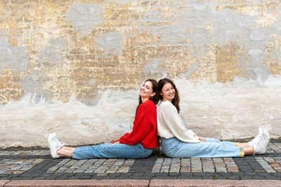 Two young women sitting back to back on the pavement by an old wall