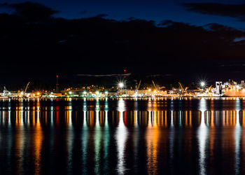 Lake by illuminated harbor against cloudy sky at night