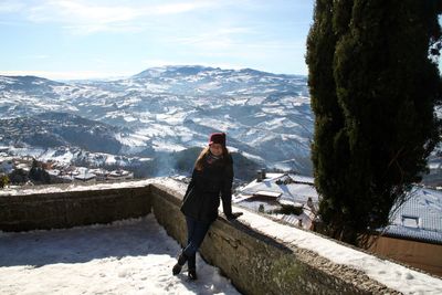 Portrait of smiling woman standing by retaining wall against snowcapped mountain