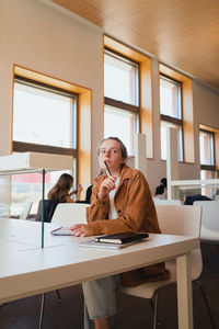 Low angle of pensive young female student touching lips with pen and looking away while sitting at table and doing homework assignment in light library