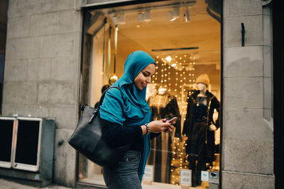 Smiling young woman using smart phone walking by store window in city