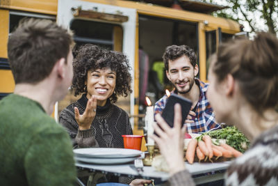 Smiling woman showing mobile phone to friends sitting at table against caravan