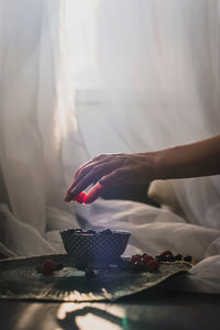 A woman's hand holding a raspberry. underneath is a little bowl more blueberries.