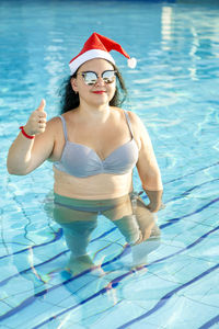 A woman in a santa hat and sunglasses and a swimsuit in the pool on vacation shows her class 