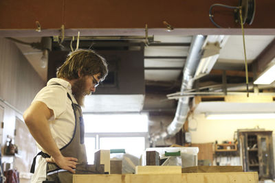 Side view of carpenter with hand on hip standing at workbench in workshop