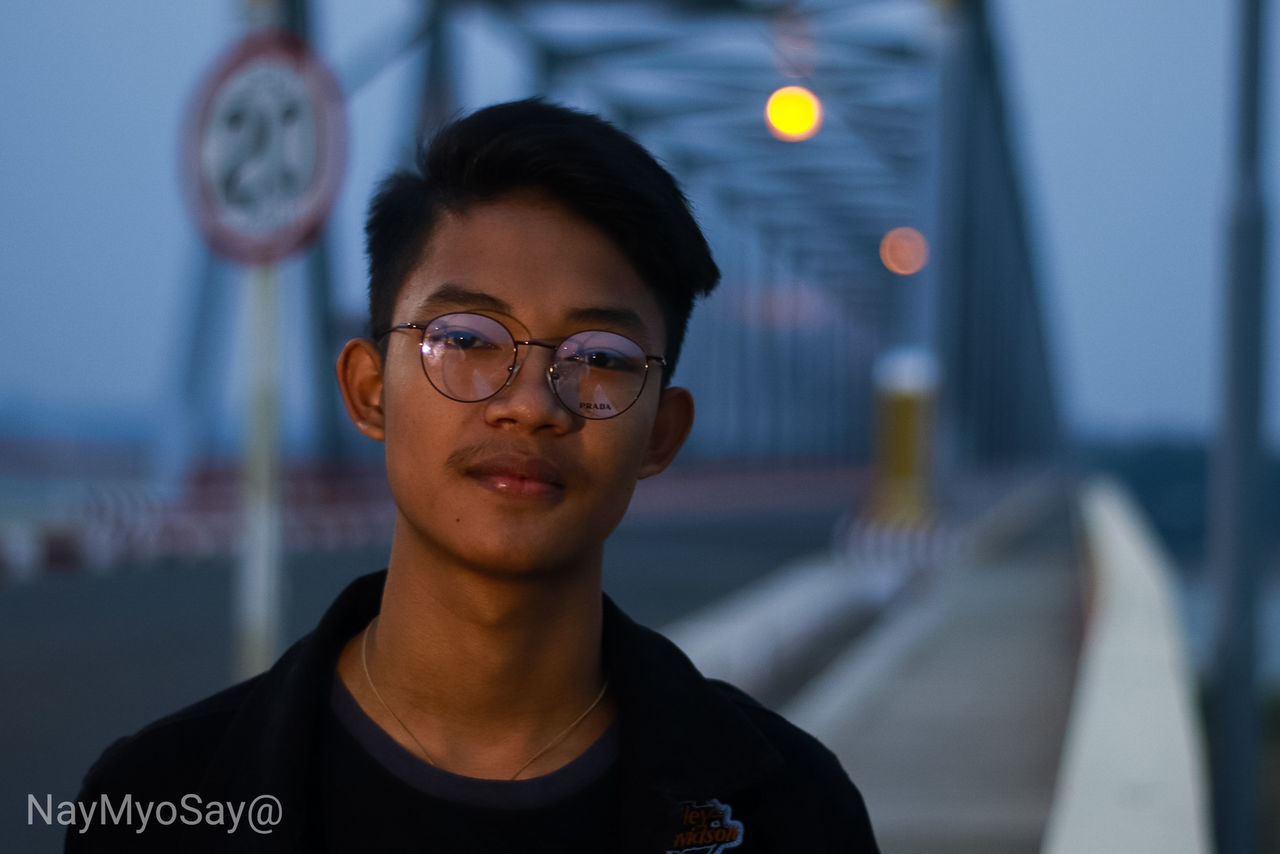 one person, front view, glasses, eyeglasses, headshot, young adult, focus on foreground, real people, lifestyles, portrait, leisure activity, young women, standing, casual clothing, smiling, looking at camera, adult, illuminated, outdoors, contemplation