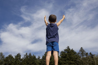 Rear view of boy with arms raised standing against sky