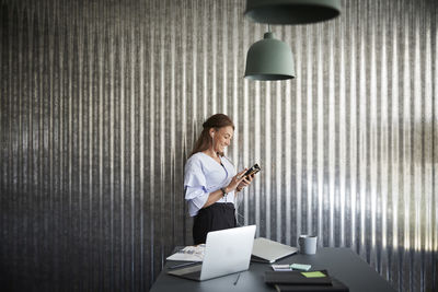 Smiling mature businesswoman using mobile phone while standing against corrugated iron wall at creative office