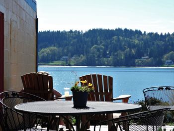 Empty chairs and table by lake against sky