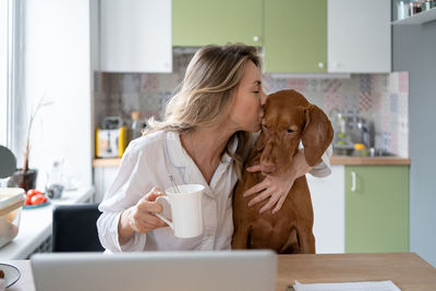 Woman in sleepwear kissing hugging her dog, sitting on chair in kitchen room, holding mug of tea. 