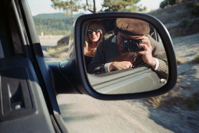 Side-view mirror with reflection of man photographing while sitting with woman in car