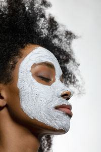 Young woman with eyes closed and facial mask on face against white background