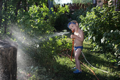 Happy little boy in watersport goggles with a pistol in hands watering the garden in summer cottage