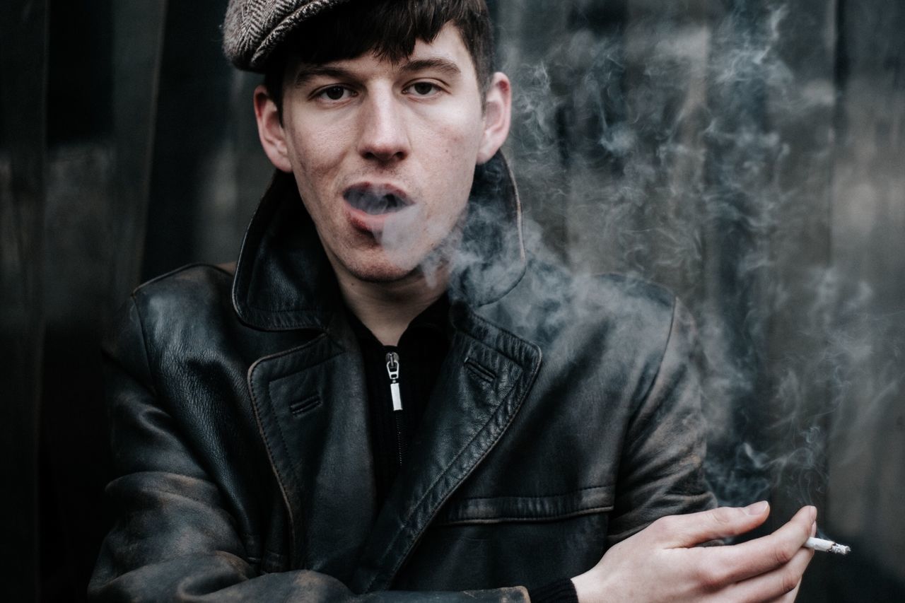 smoke - physical structure, one person, portrait, front view, smoking - activity, young adult, smoking issues, social issues, men, indoors, adult, real people, tobacco product, young men, clothing, bad habit, leather jacket