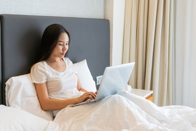 Asian girl working on a laptop while sitting on bed at home. technology and lifestyle concept. 