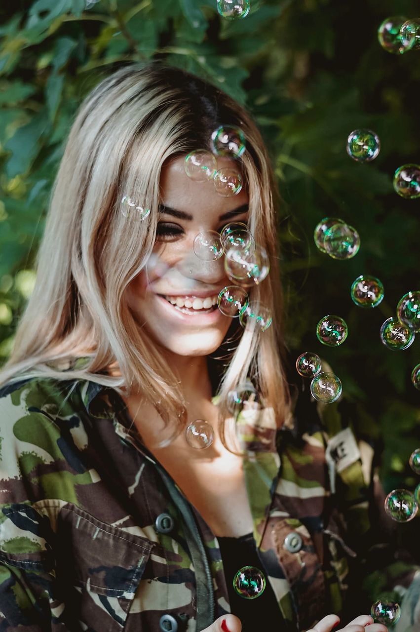 one person, smiling, happiness, women, portrait, bubble, adult, emotion, long hair, enjoyment, nature, cheerful, bubble wand, hairstyle, fun, young adult, headshot, fragility, leisure activity, plant, childhood, child, outdoors, lifestyles, female, day, blowing, smile, blond hair, teeth, carefree, liquid bubble, holding, tree, person, photo shoot, looking, front view, standing, joy, flower, waist up