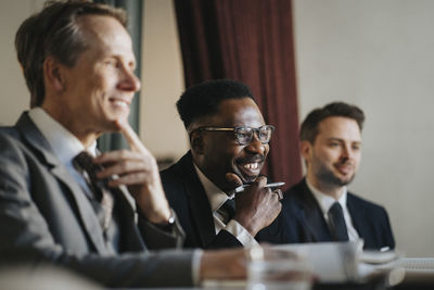 Smiling businessman with hand on chin sitting with male colleagues during meeting at office