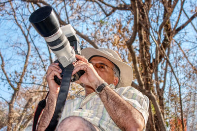 Low angle view of mature man holding camera by tree