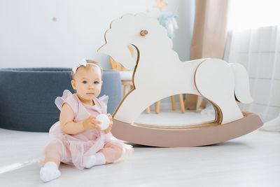 A cute baby in a pink dress holds a small white ball in her hands and sits on the floor