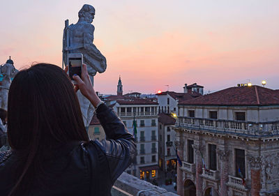 Rear view of woman photographing statue and buildings at sunset
