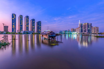 Illuminated buildings by lake against sky in city at dusk