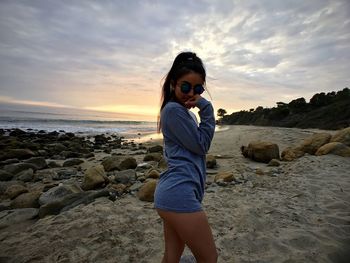 Side view of sensuous woman standing at sandy beach against cloudy sky