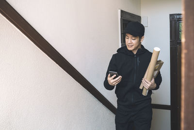 Smiling delivery man with package using phone against wall