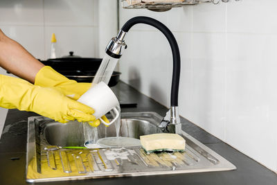 Close up human hands in yellow rubber gloves washing a ceramic coffee mug in the kitchen sink.