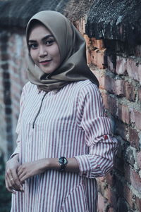 Portrait of smiling young woman wearing hijab while standing against brick wall