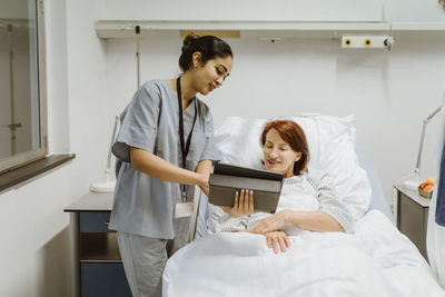 Smiling nurse showing digital tablet to senior female patient lying on bed in hospital